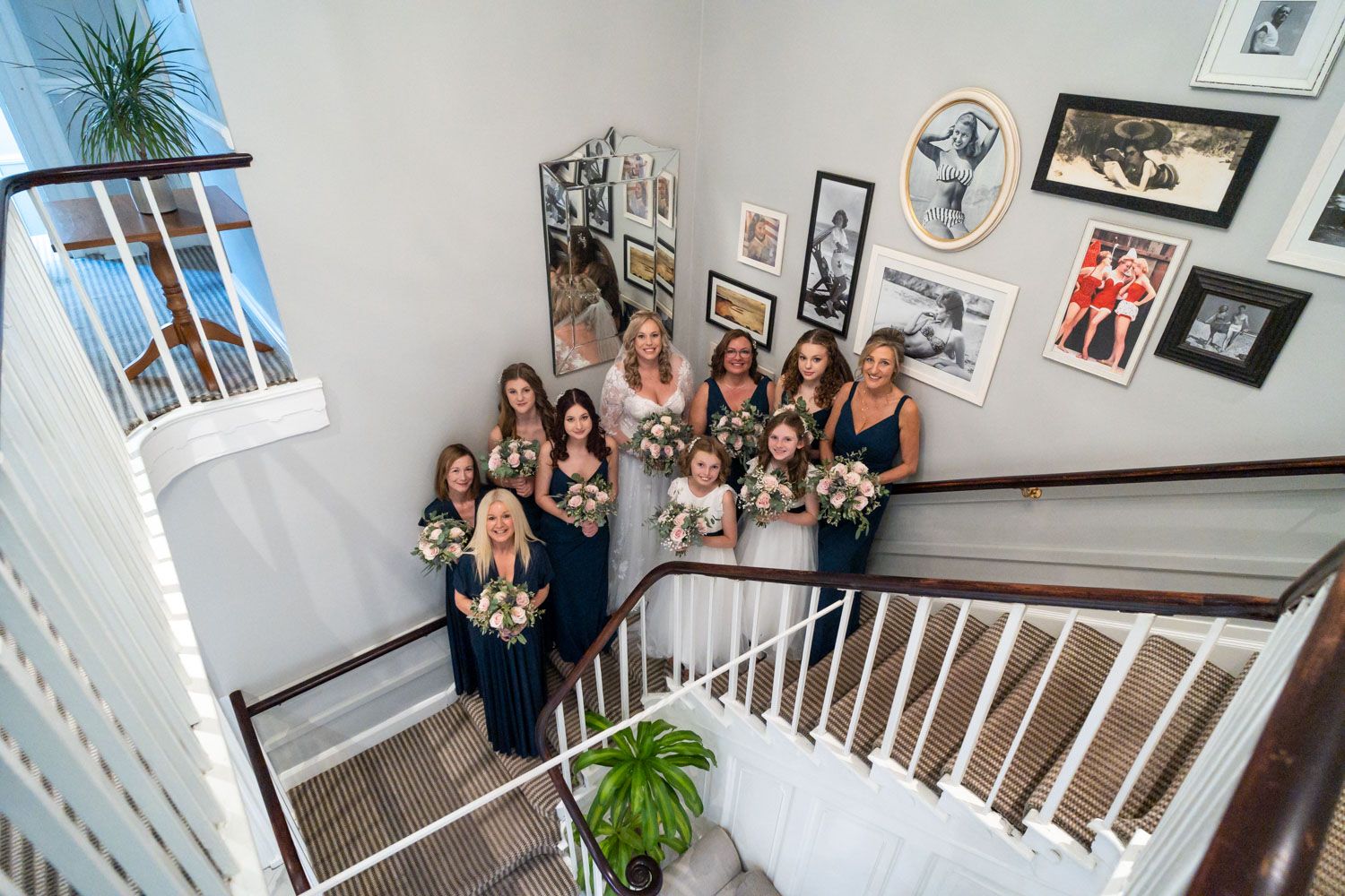 A group of elegantly dressed wedding attendees pose together on a white staircase near a photo-decorated wall.