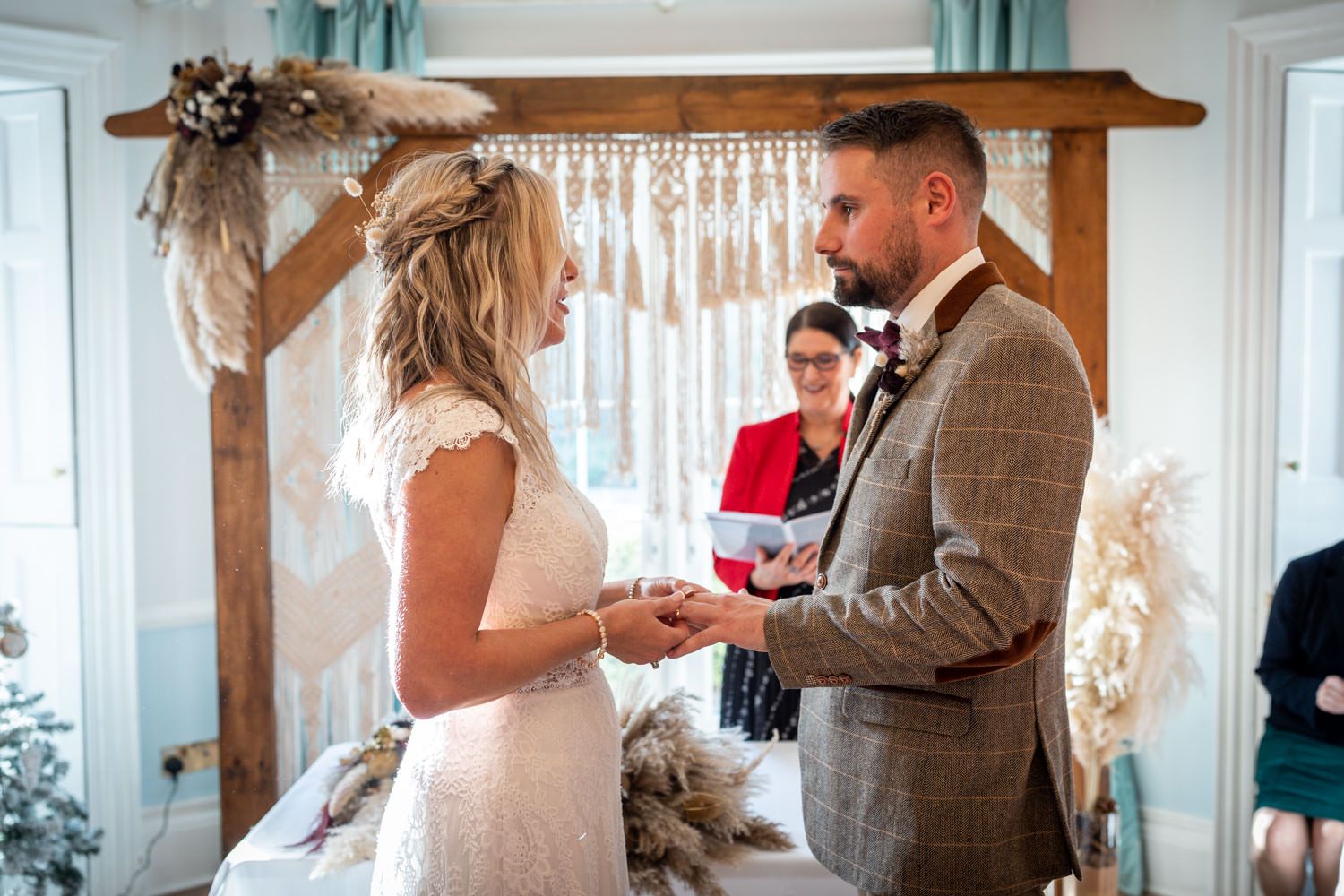 A couple exchanges wedding rings during an intimate indoor ceremony under a wooden arch with macrame decor.