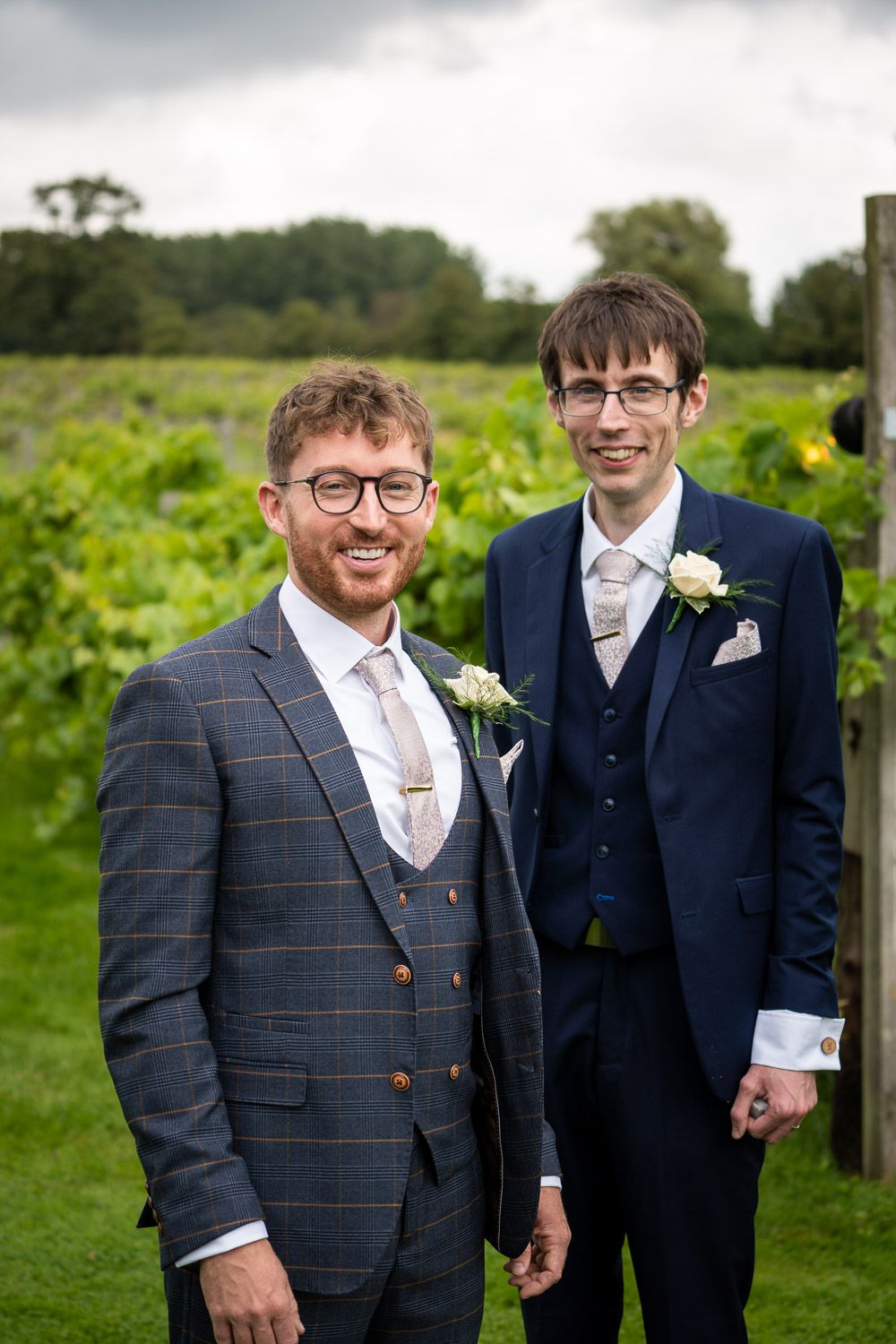 Two men in formal suits standing together outdoors in front of a green vineyard on an overcast day.