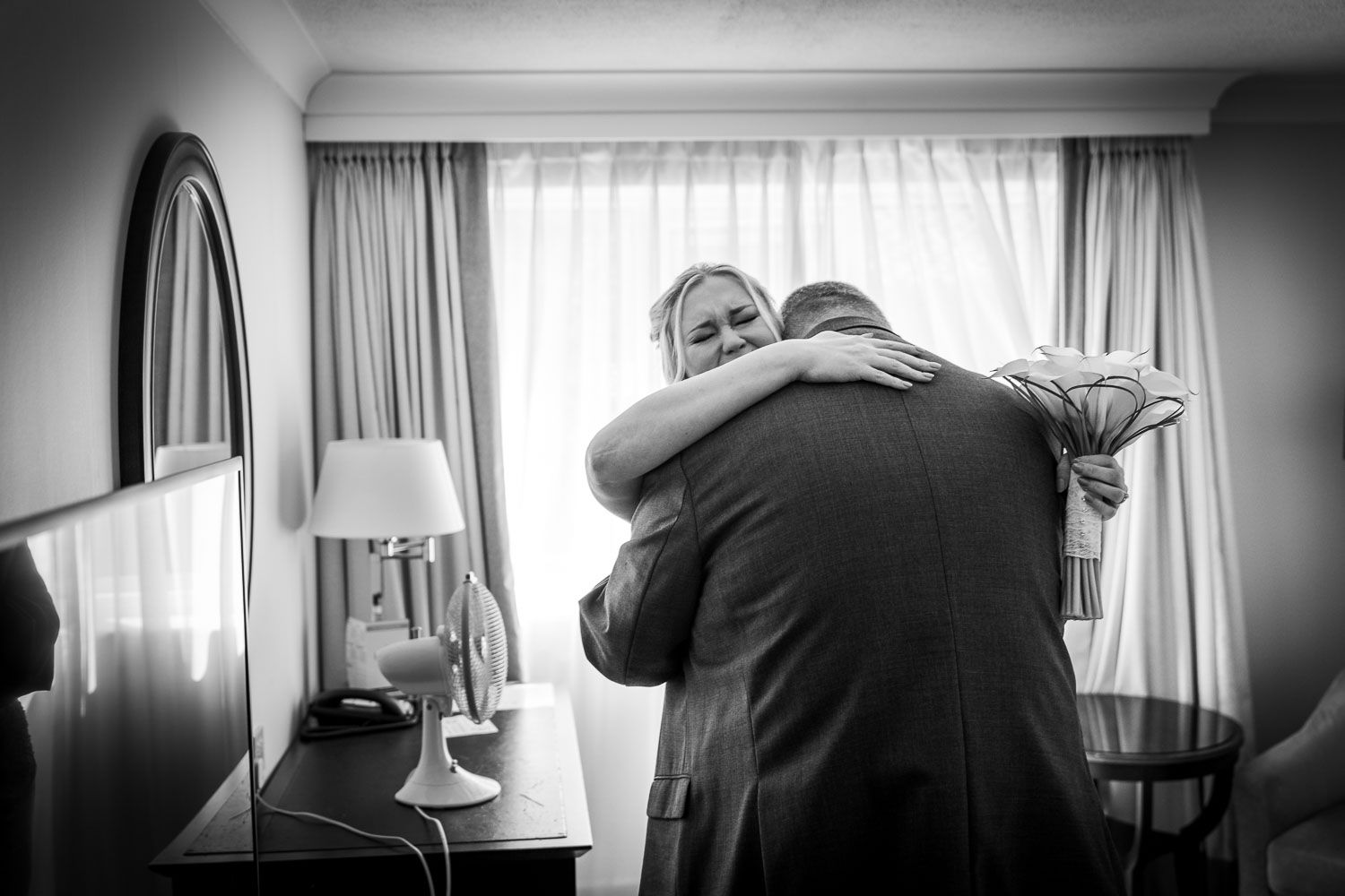 An emotional embrace in black and white photograph taken in a hotel room with curtained windows and a table lamp.