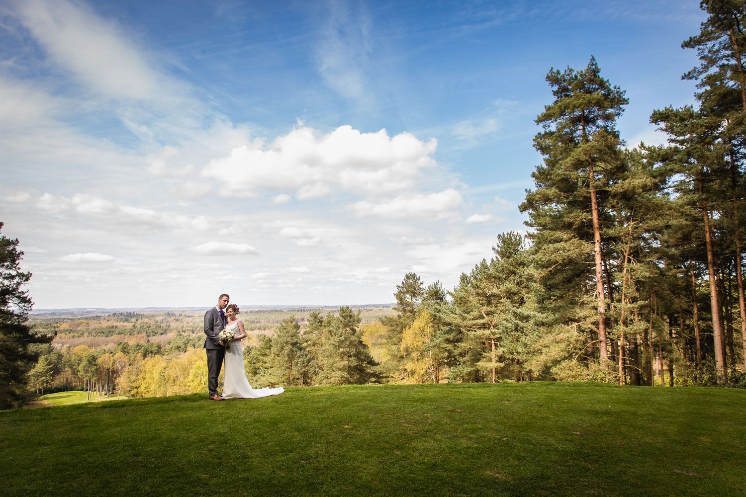 A couple embraces on a hilltop overlooking a scenic autumn landscape with tall pine trees and blue skies.