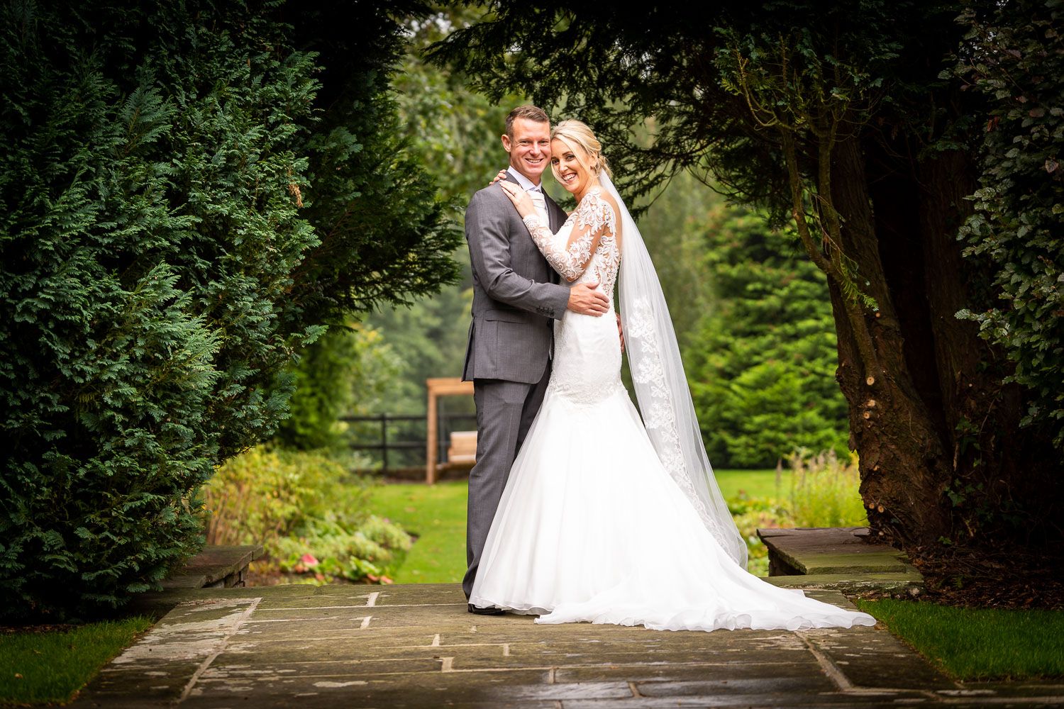 Bride and groom embracing on garden pathway surrounded by lush green hedges and trees.