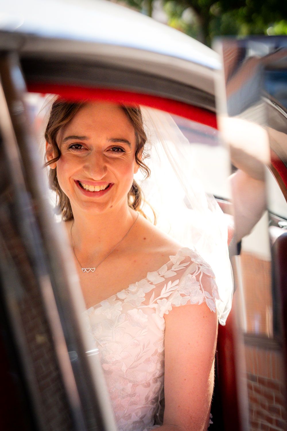 Smiling bride in lace wedding dress and veil photographed through a car window on wedding day.