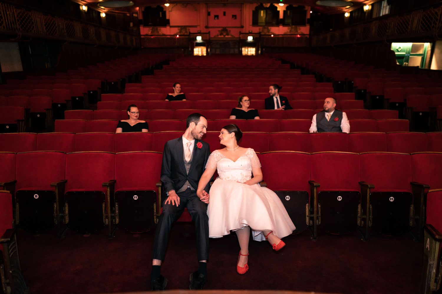 Bride and groom sitting together in a vintage red theater with wedding guests seated behind them.
