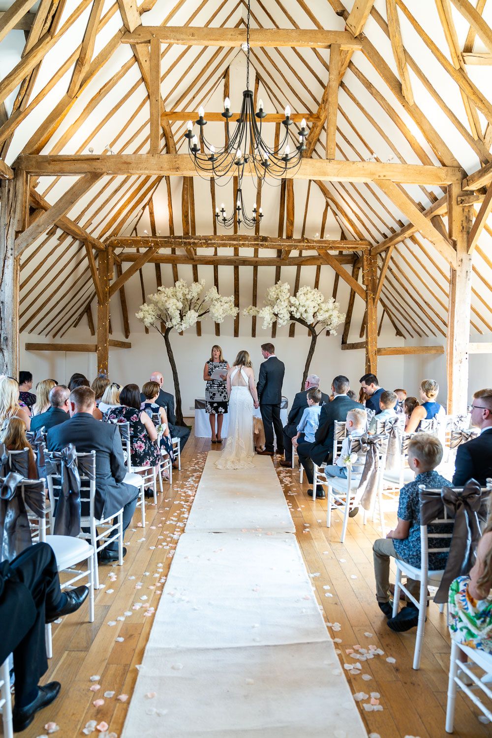 Indoor rustic barn wedding ceremony with exposed wooden beams, white decorations and chandelier in a vaulted ceiling venue.
