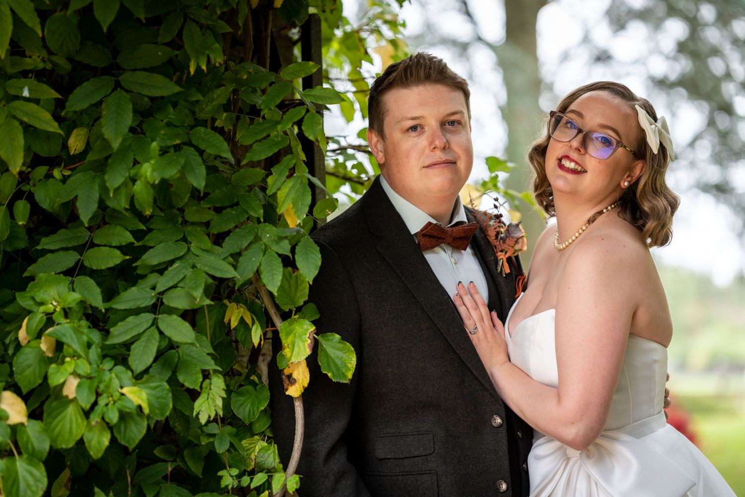 A couple poses outdoors on their wedding day next to green foliage in formal attire.