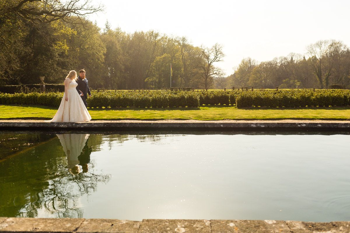 A stunning reflection appears in a pond at sunset as people in wedding attire stand at the water's edge in a manicured garden.