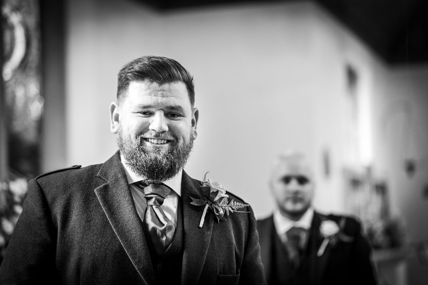 Joyful groom in a black suit and tie smiles during wedding ceremony while person stands in background.