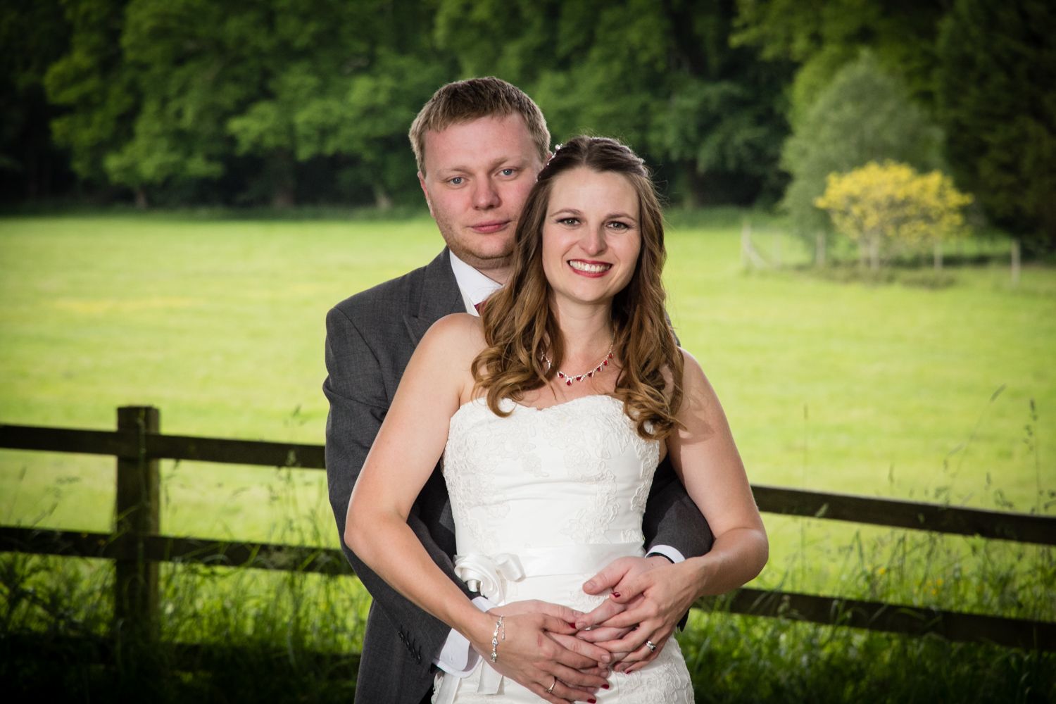 Couple poses together in formal wear by a wooden fence with lush green landscape in the background.