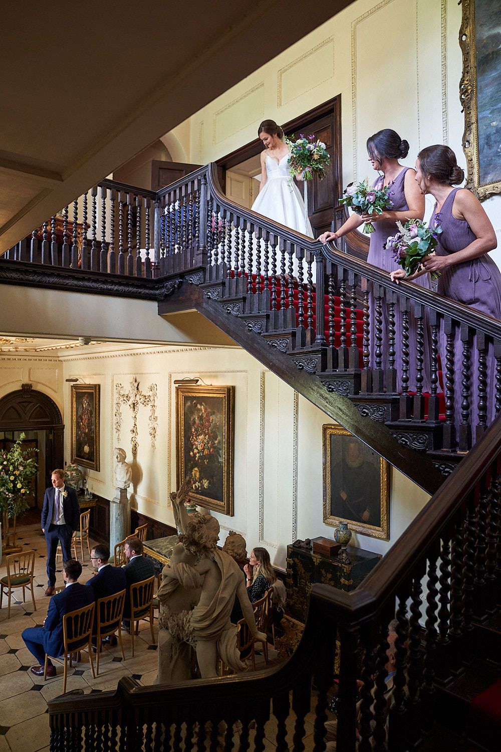 Wedding party members descend an elegant staircase with ornate railings and decorative art in a grand historic venue.