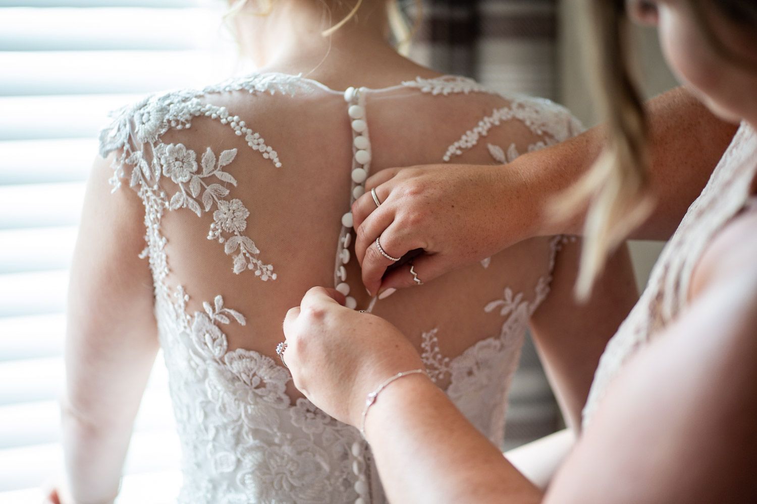 Wedding dress detail showing delicate white lace pattern and buttons being fastened on the back.