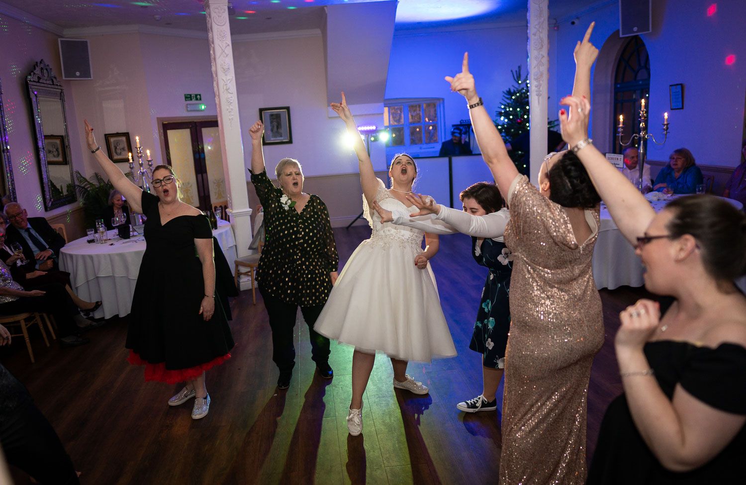 Wedding party guests dancing and celebrating on a dance floor illuminated by blue lighting at a reception venue.