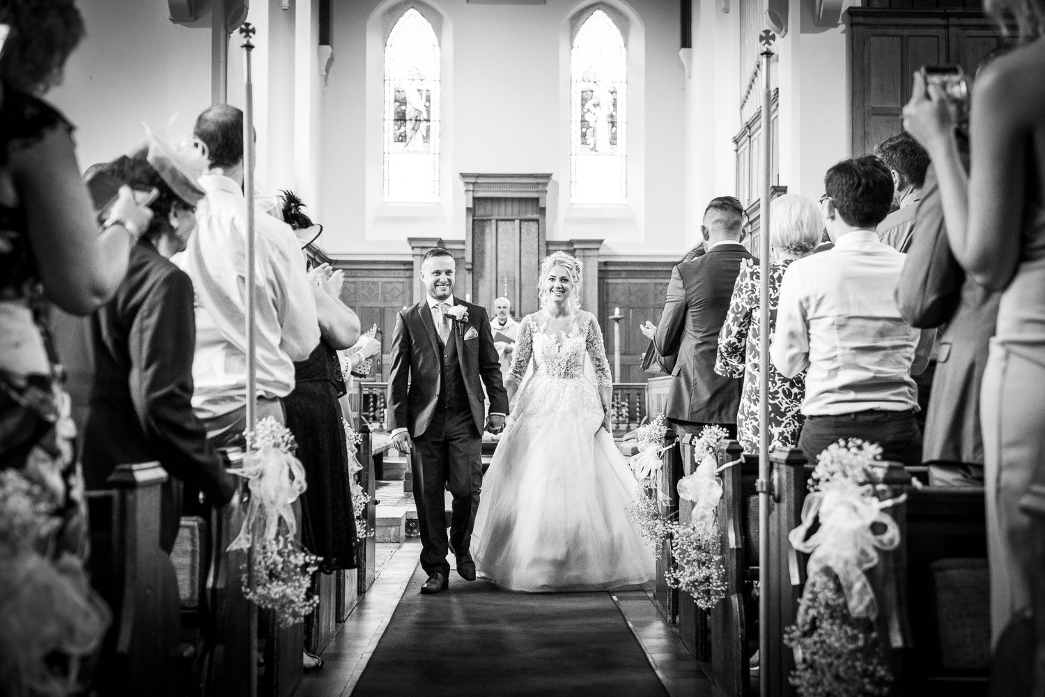 A black and white photograph shows a couple walking down a church aisle decorated with baby's breath flowers.