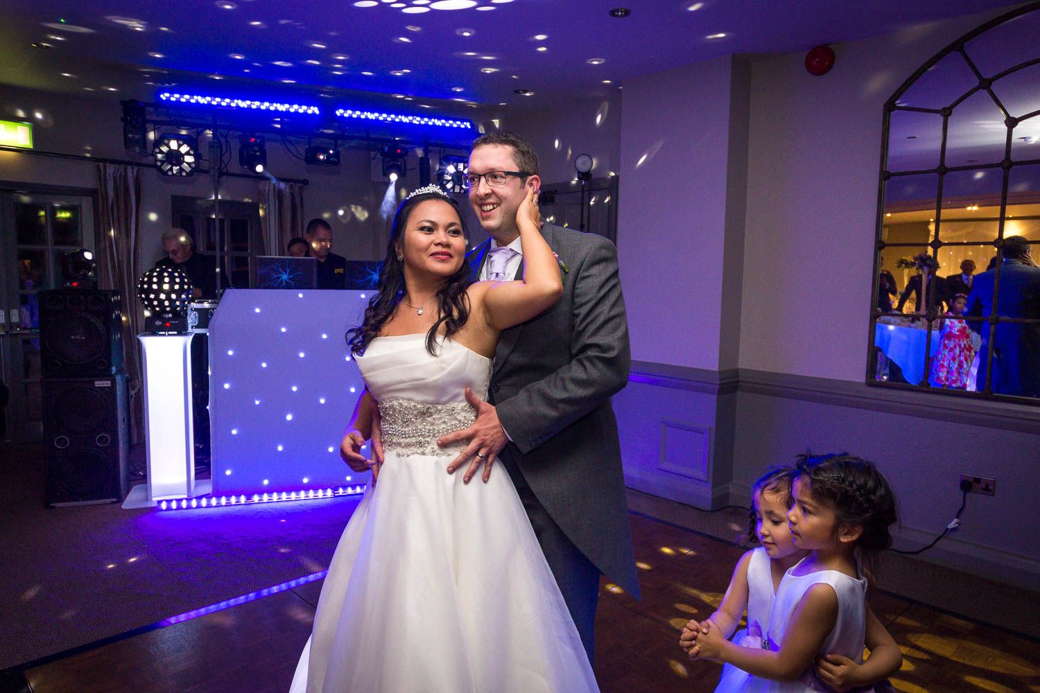 Couple dances at wedding reception with blue mood lighting and illuminated dance floor as young guest watches.