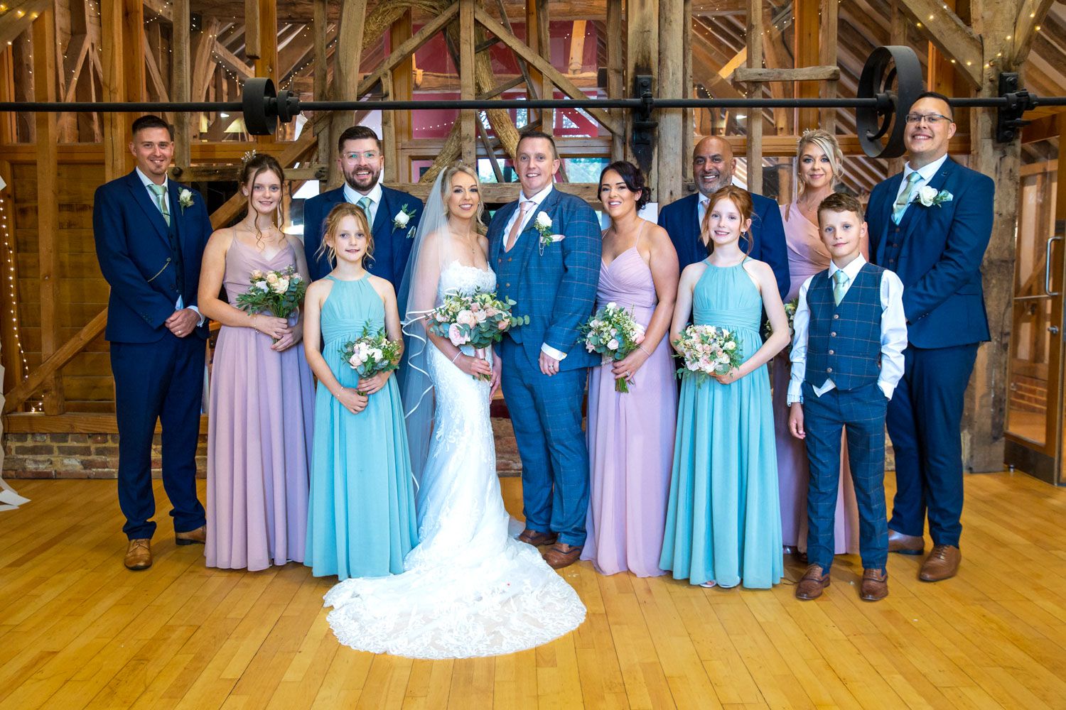 Wedding party members lined up in formal attire with blue and pink dresses in a rustic wooden venue.