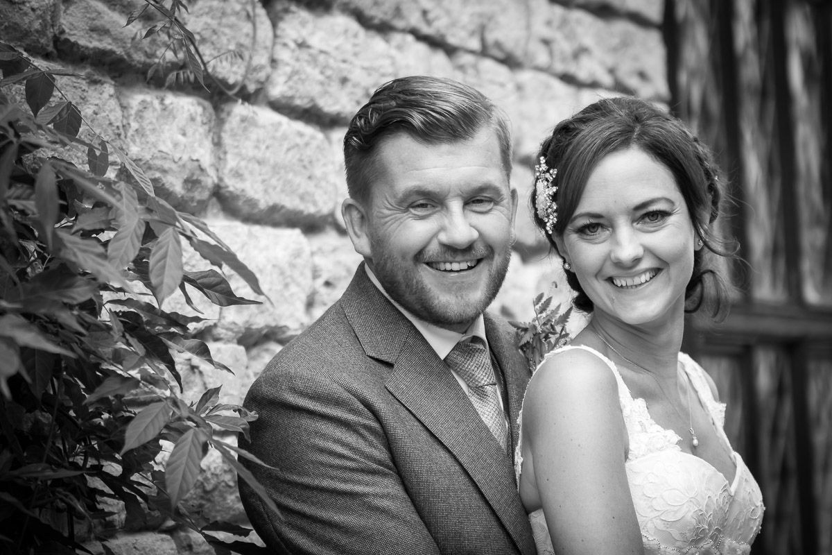 A joyful black and white wedding portrait of a couple posing together against a stone wall outdoors.