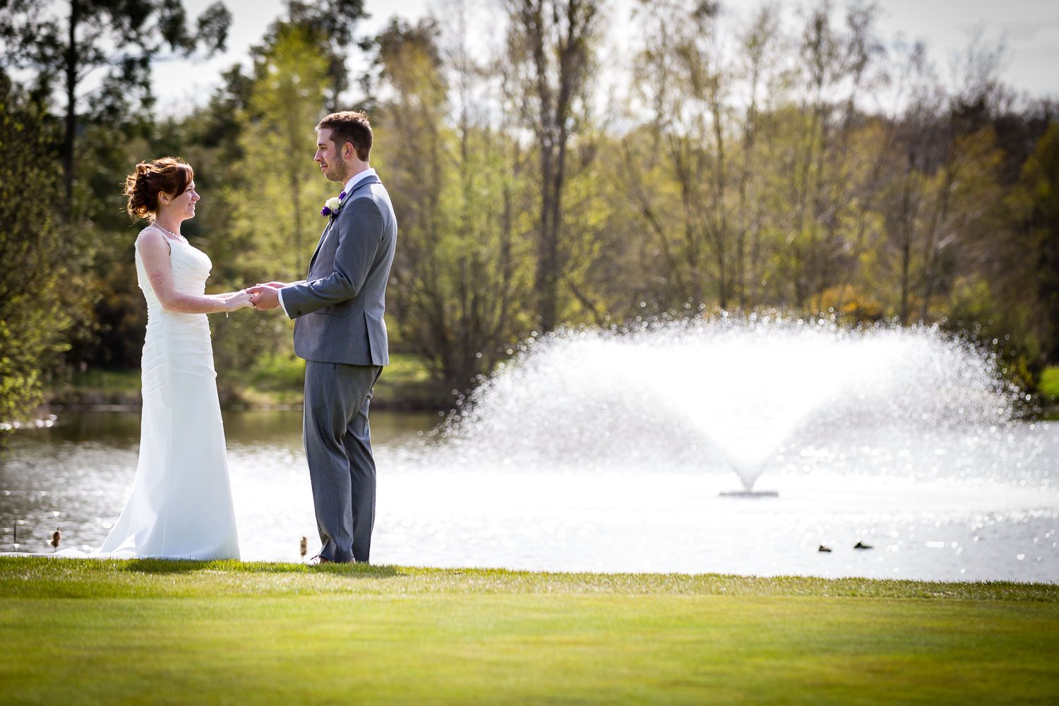 A wedding couple stands on a lush golf course with a beautiful fountain spraying water in the background.