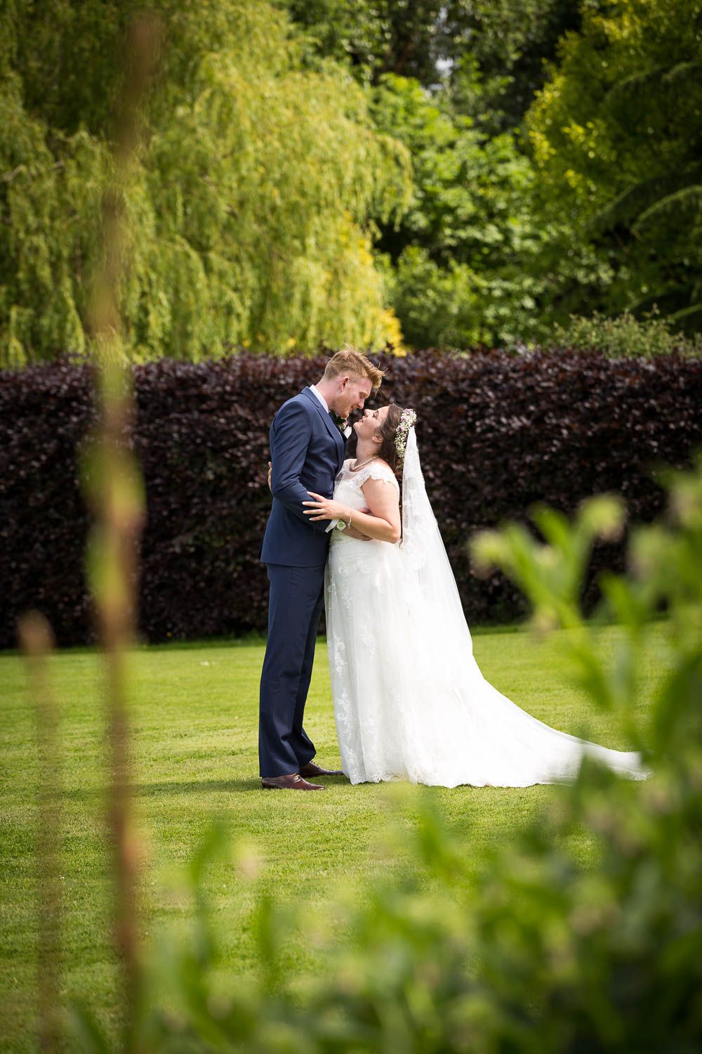 A romantic garden wedding scene with a couple embracing on a lush lawn with weeping willows and hedges in the background.