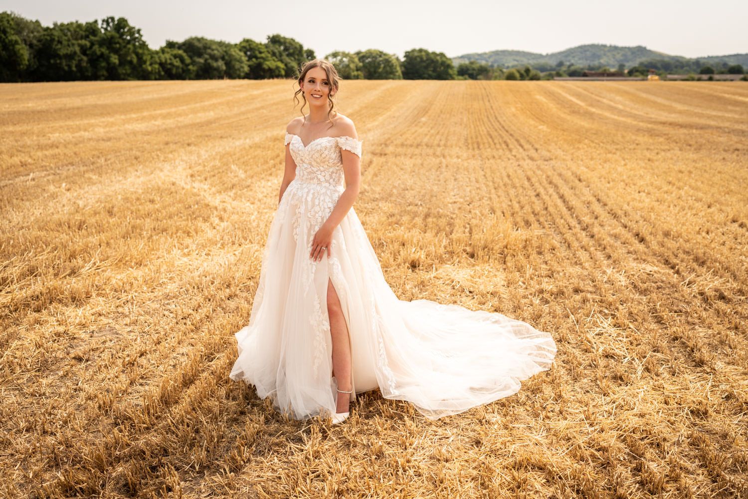 A stunning wedding dress with flowing train photographed against a golden harvested field at sunset.