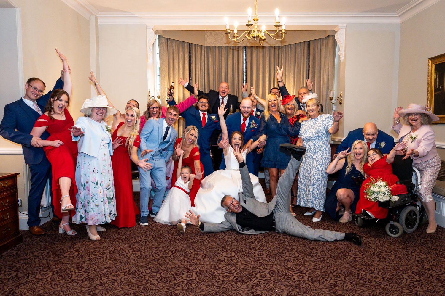 Large group of wedding guests pose enthusiastically together for a fun group photo in an elegant room with chandelier.