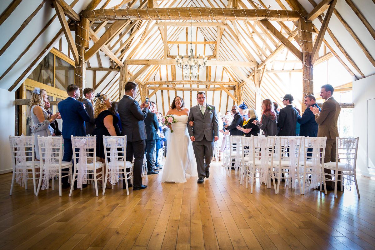 Wedding ceremony taking place in a rustic wooden barn venue with exposed beams and white chairs lined up for guests.