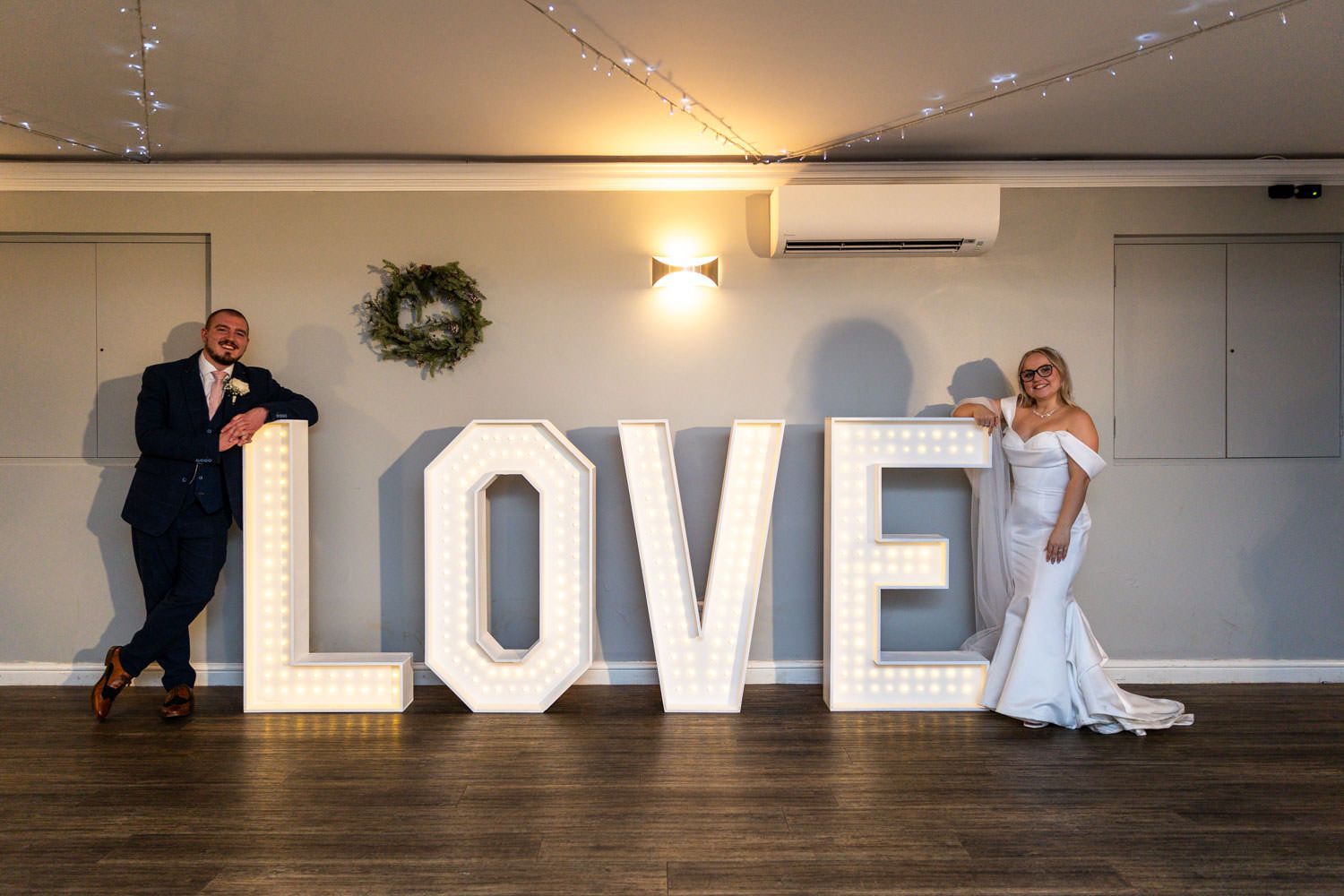 Newlywed couple poses with illuminated LOVE marquee letters at wedding reception on wooden floor.