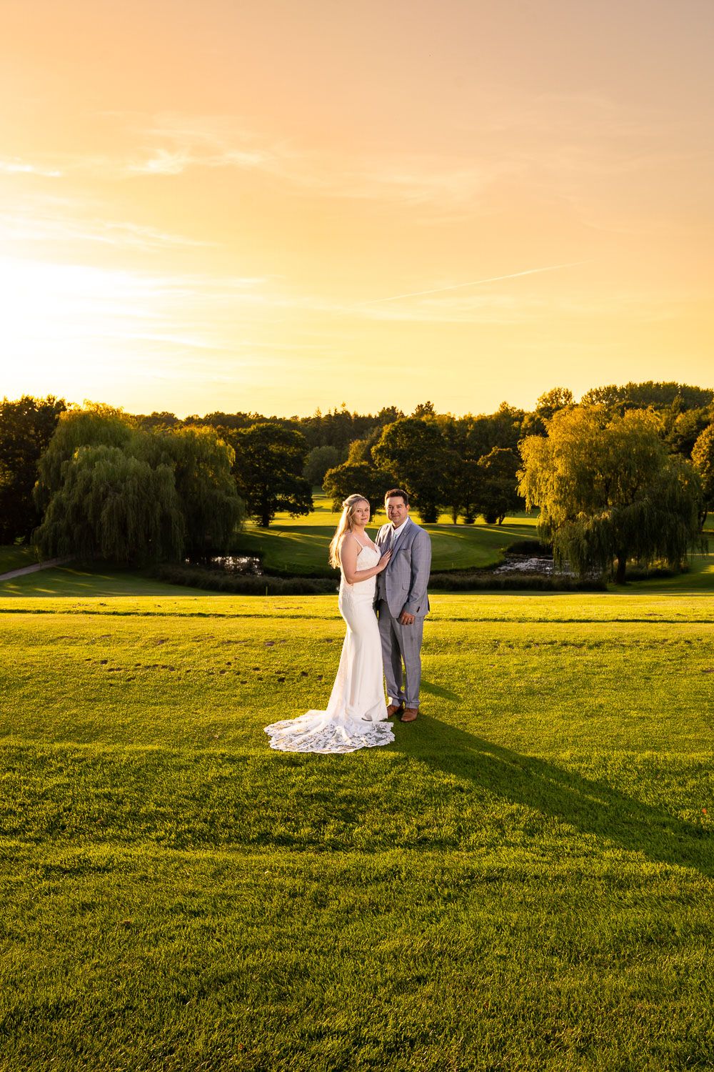A golden hour wedding portrait showing a couple on a sweeping lawn with willow trees silhouetted against the sky.