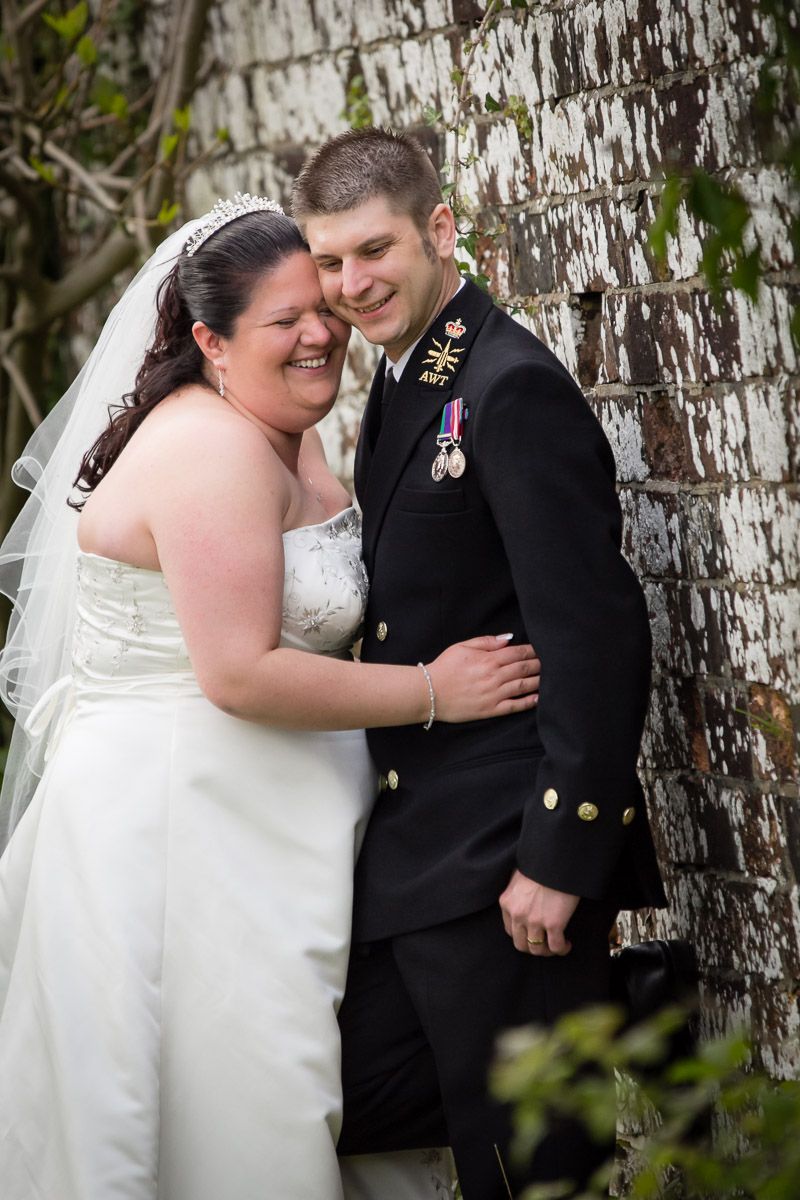 A couple share a joyful moment on their wedding day against a rustic white brick wall.