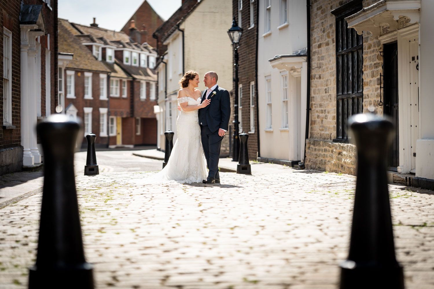A romantic couple in wedding attire dances together on a cobblestone street between historic buildings and black bollards.