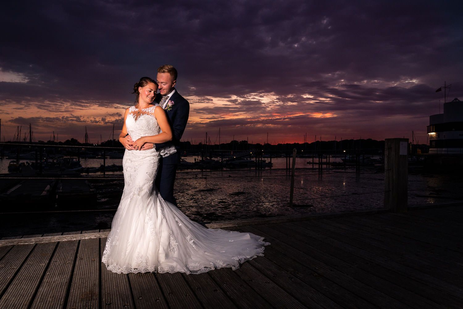 Bride and groom posing romantically on marina dock beneath dramatic purple and orange sunset sky.