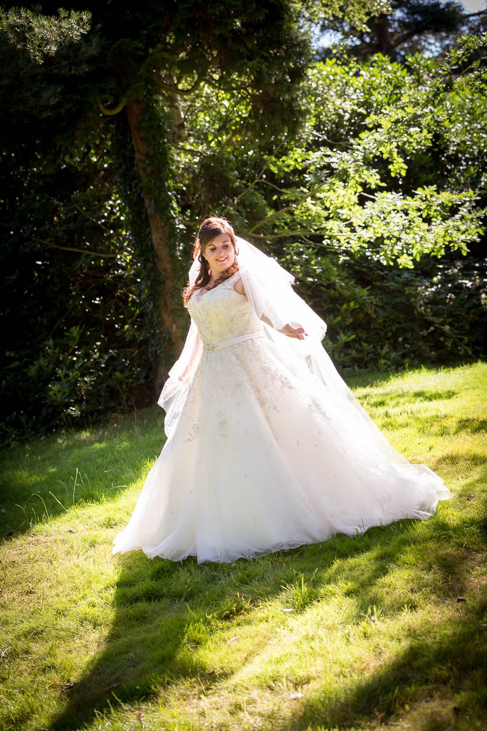 A wedding gown with flowing tulle fabric twirls in dappled sunlight on a grassy lawn surrounded by green trees.