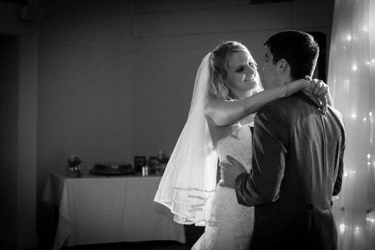 An intimate black and white photo of newlyweds sharing their first dance under twinkling lights.
