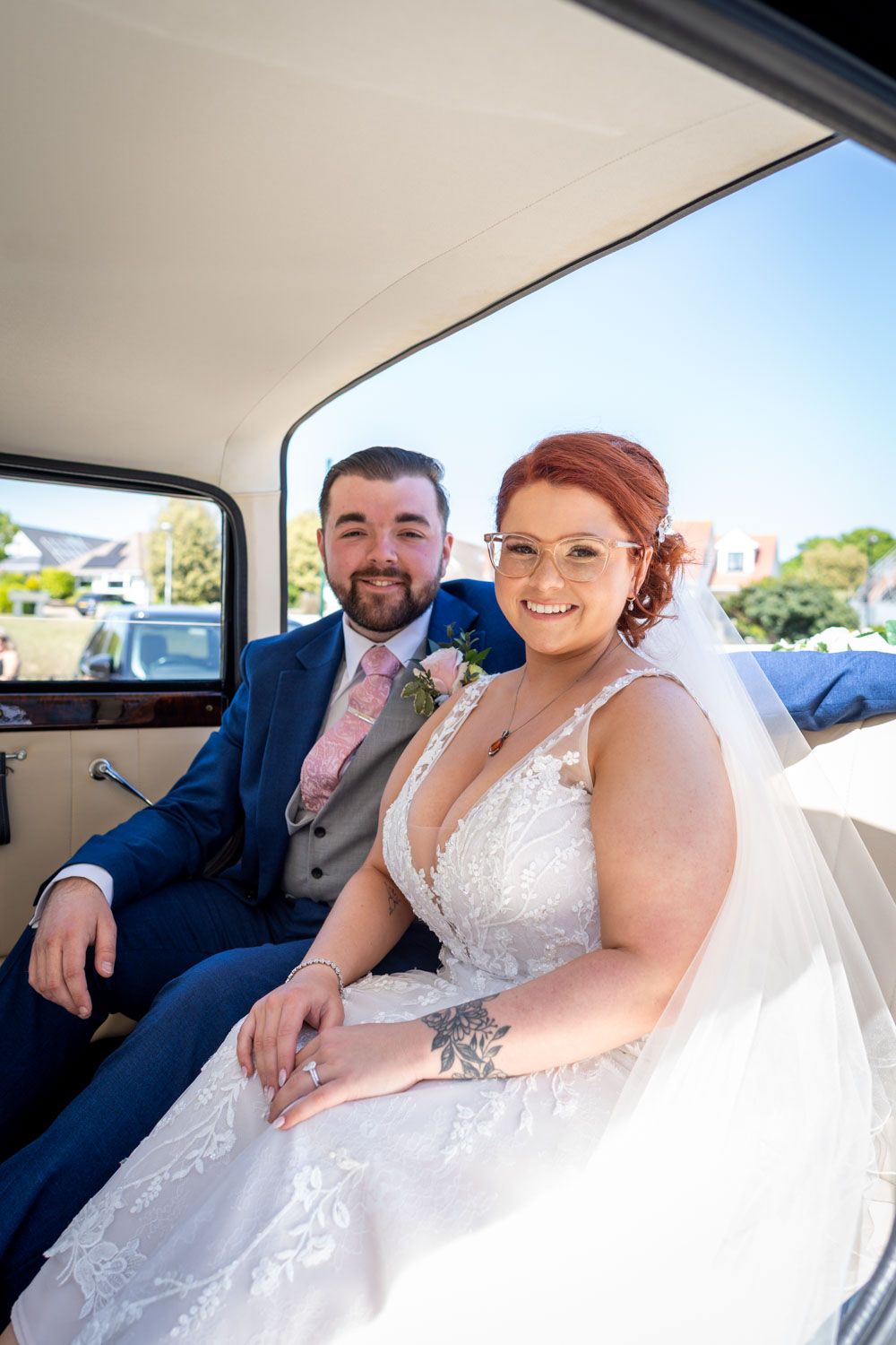 Newlywed couple smiling while sitting together in a classic car on their wedding day.