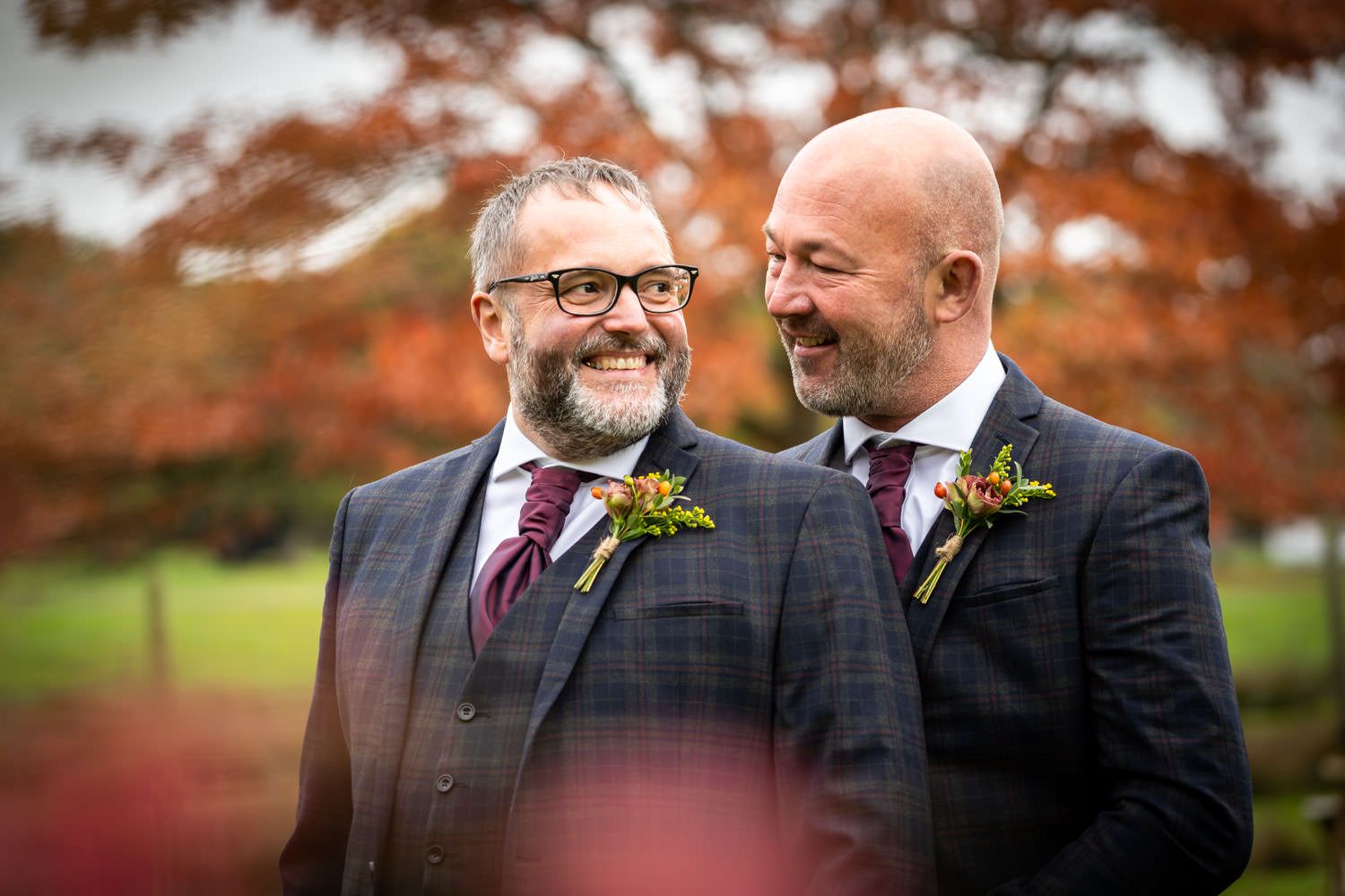 Two grooms in matching plaid suits smiling joyfully outdoors with autumn foliage background.