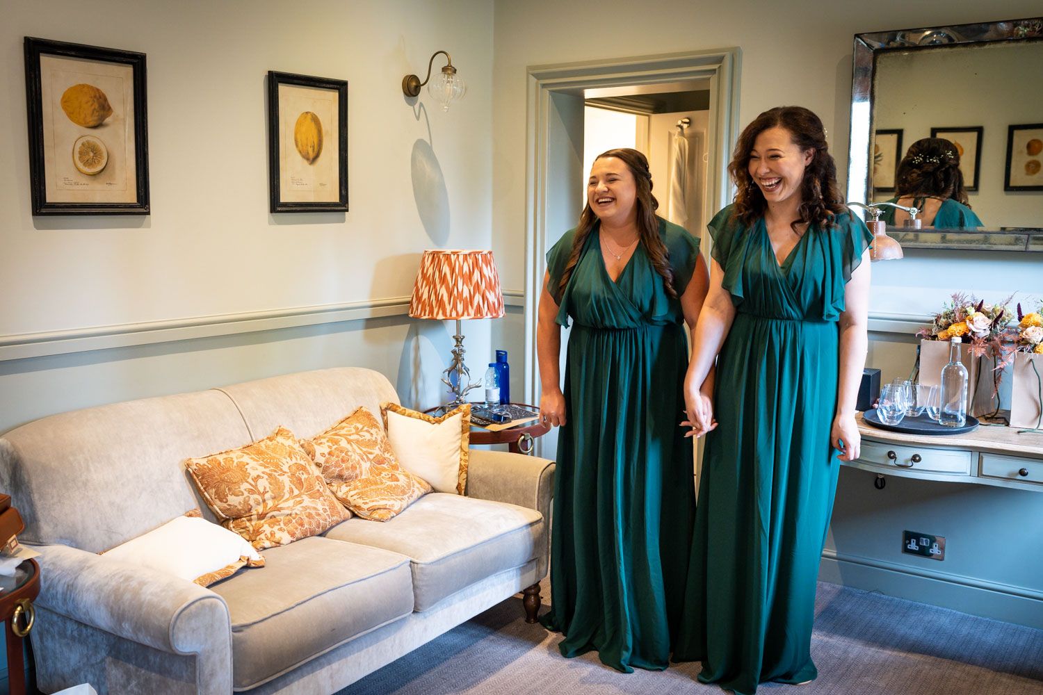 Two people in matching emerald green bridesmaid dresses laugh together in an elegant living room setting.