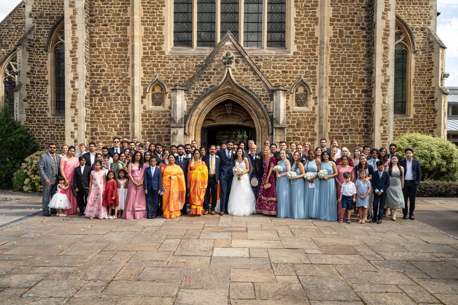 Large wedding party in colorful formal attire gathered outside historic stone church entrance for group photo.