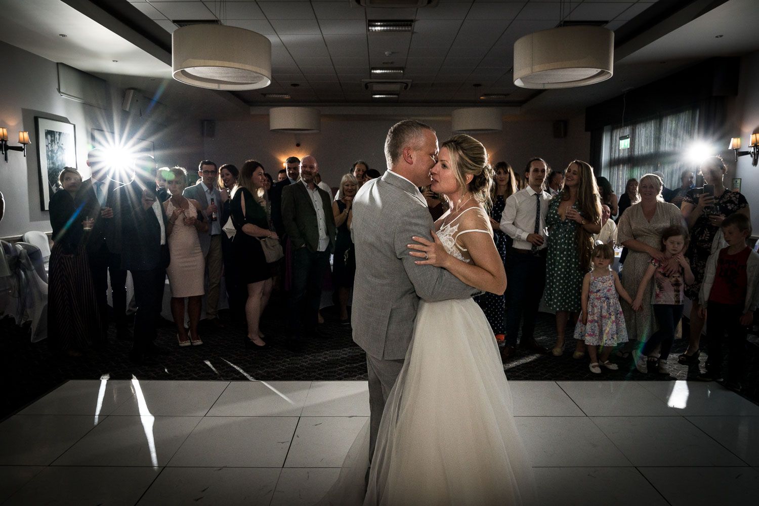 A couple shares their first dance as newlyweds on a dimly lit dance floor surrounded by wedding guests and camera flashes.