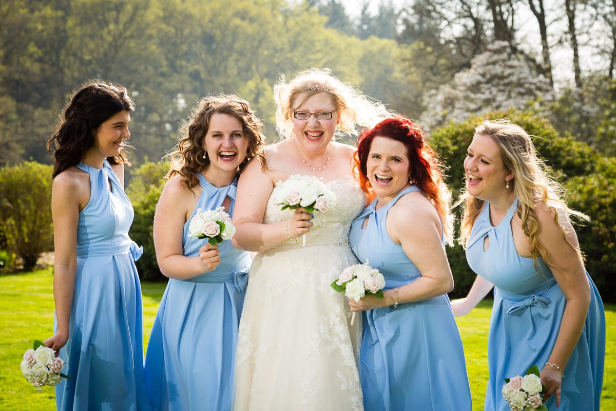 A group of people wearing light blue dresses and a person in white laugh together while holding bouquets outdoors.