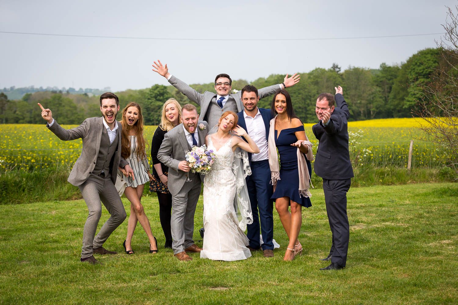 Wedding party in formal attire poses joyfully in front of a vibrant yellow rapeseed field on a green lawn.