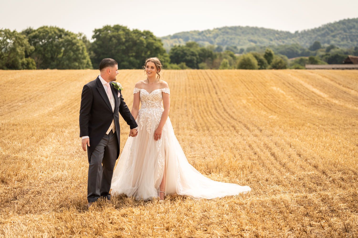 A couple in formal wedding attire stands together in a golden wheat field with rolling hills in the background.