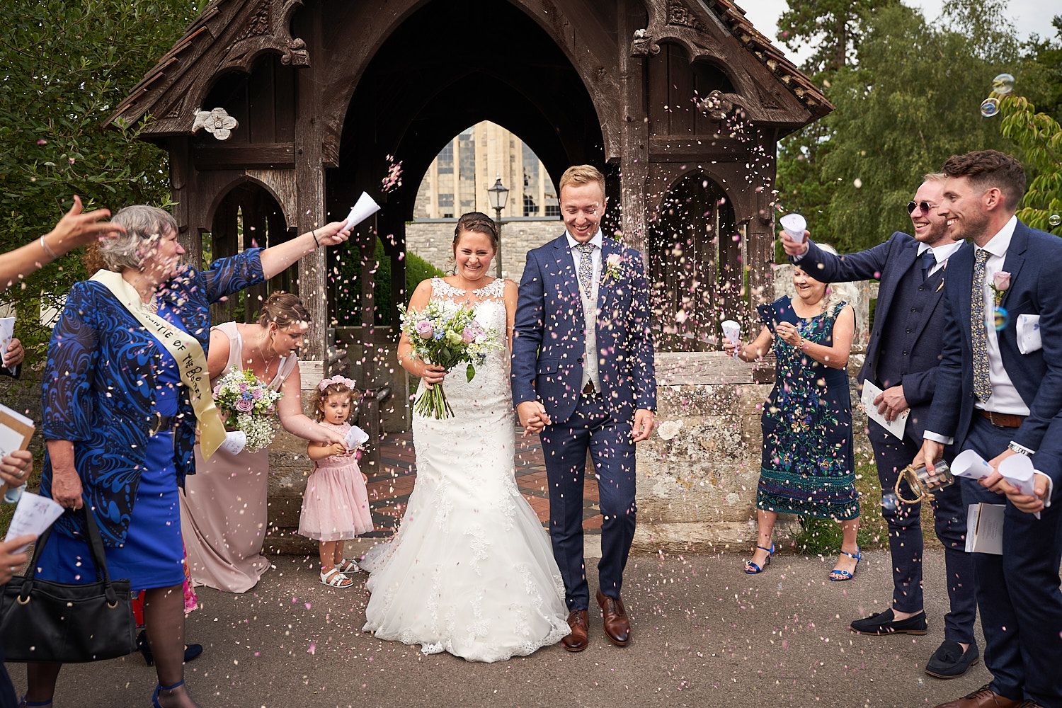 Happy couple walks under confetti shower outside rustic wooden lych gate while guests celebrate around them.