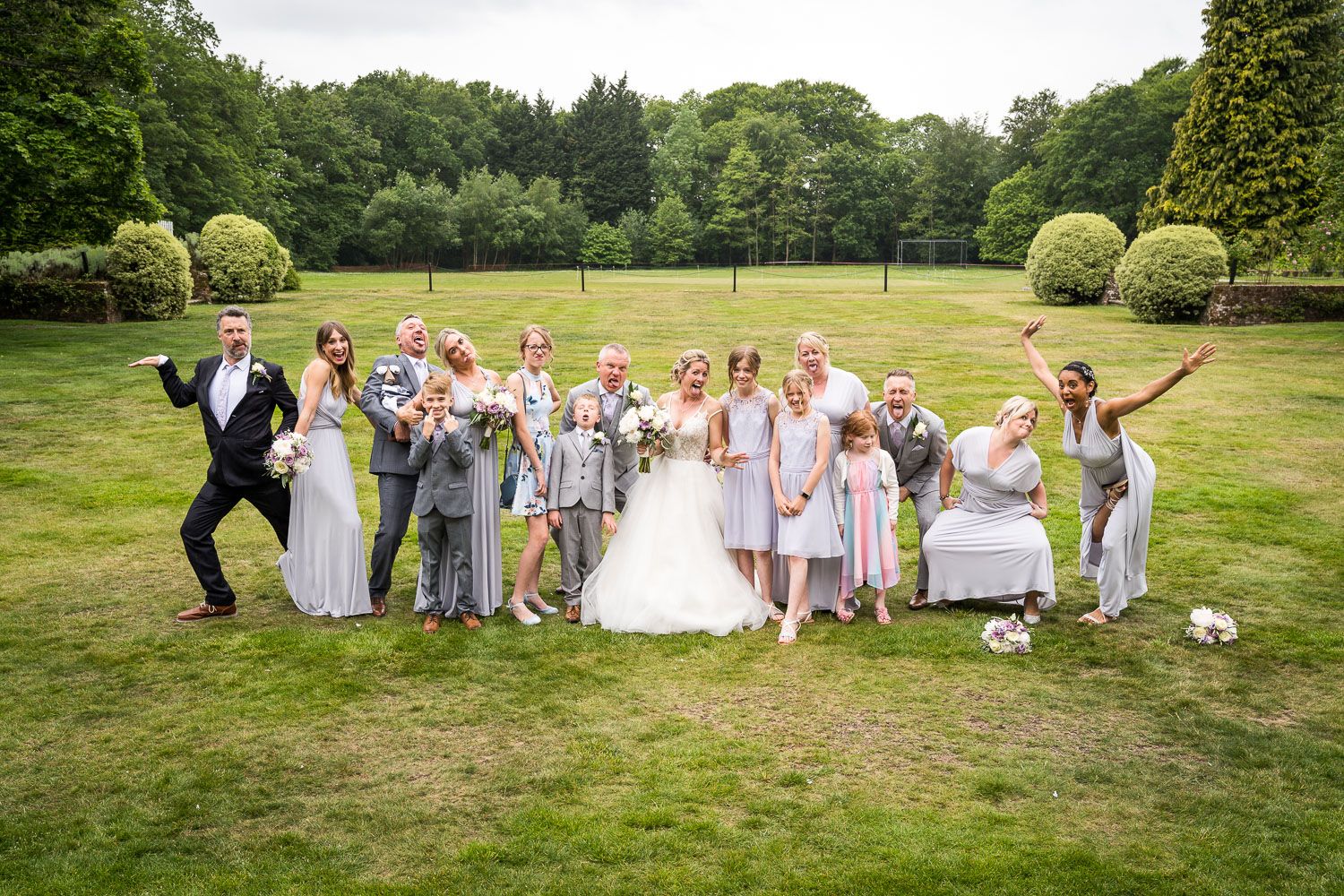 Joyful wedding party celebrates on a lush green lawn surrounded by manicured gardens and trees.