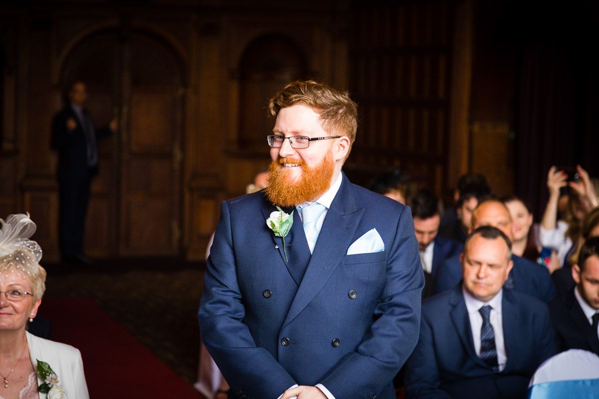 Groom wearing a navy blue suit and white floral boutonniere stands at the altar during a wedding ceremony.