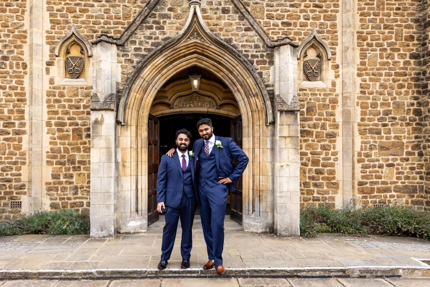 Two people in matching navy suits stand in front of an ornate stone church archway entrance.