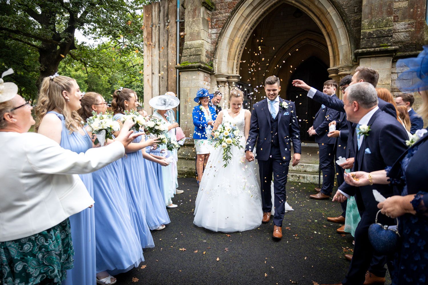 Wedding guests throwing confetti outside church entrance with bridesmaids in light blue dresses.