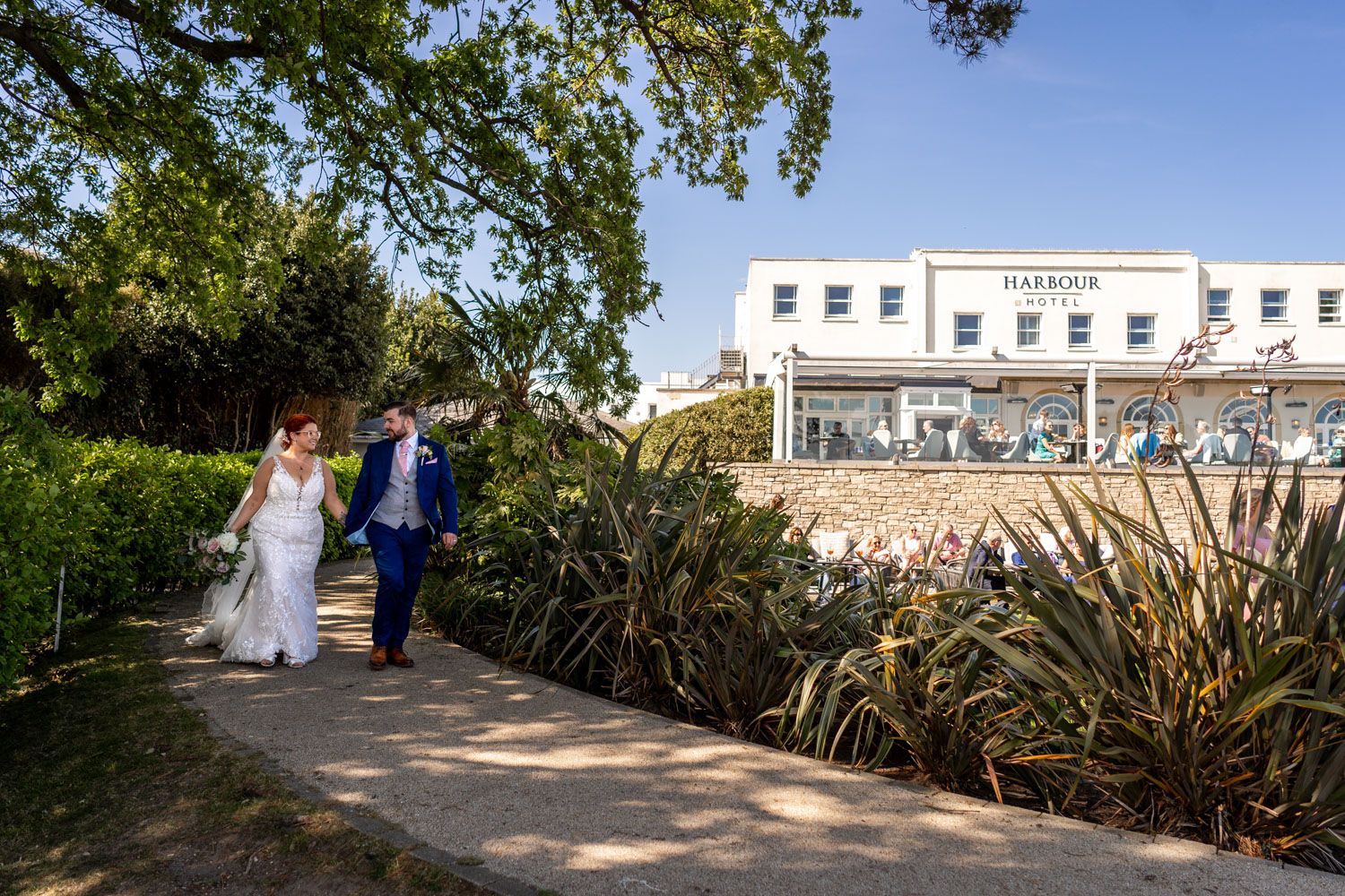 A couple walks along a garden path beside a white coastal building on a sunny day.