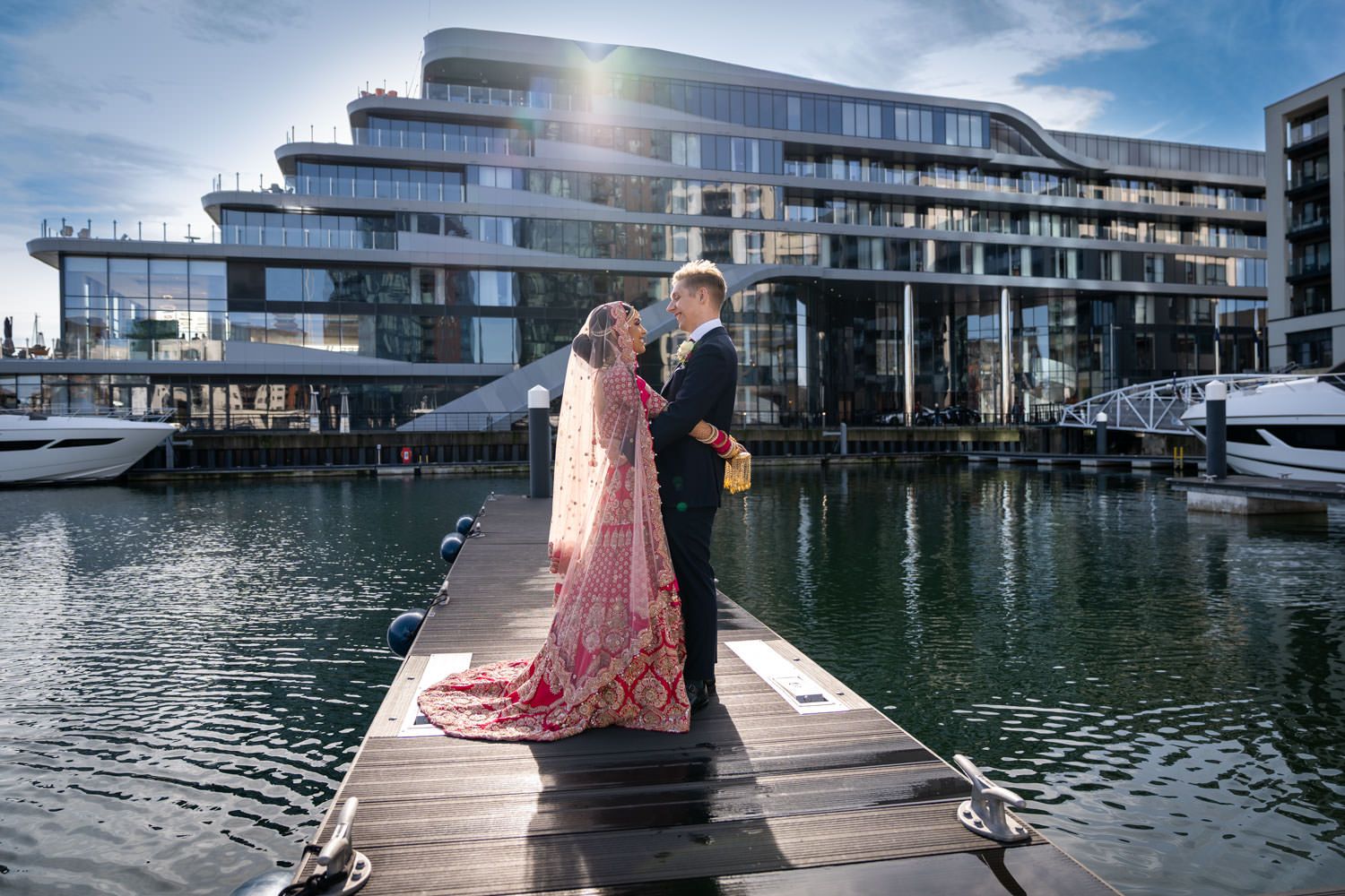 Bride in pink Indian wedding attire and groom standing on marina dock with modern waterfront building.