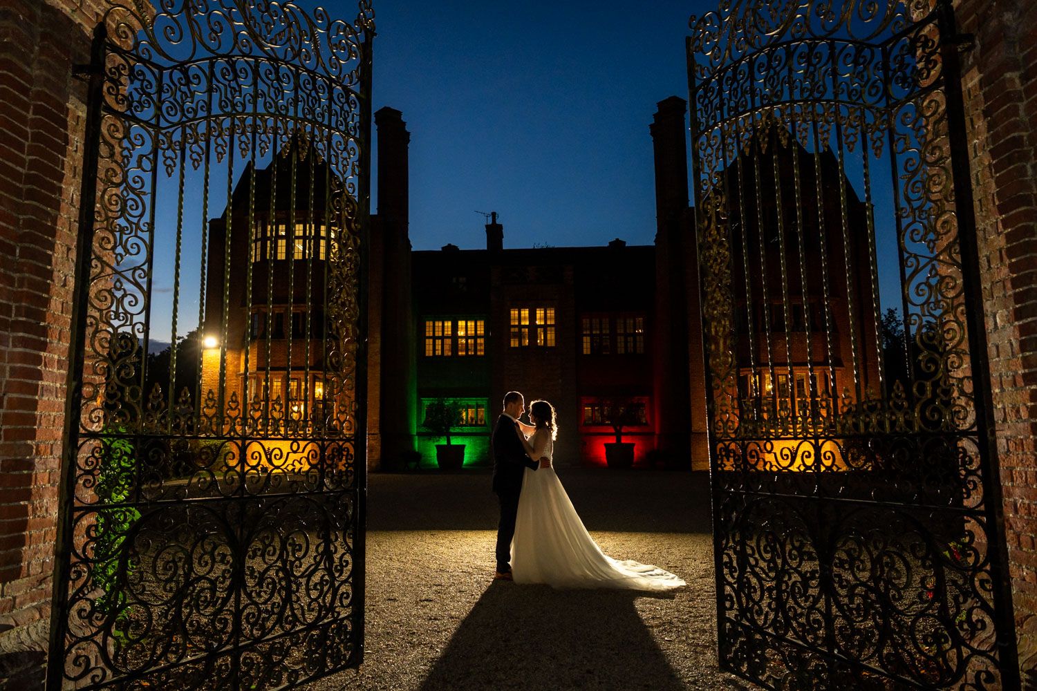 Bride and groom silhouetted at night before colorfully lit historic brick manor wedding venue.