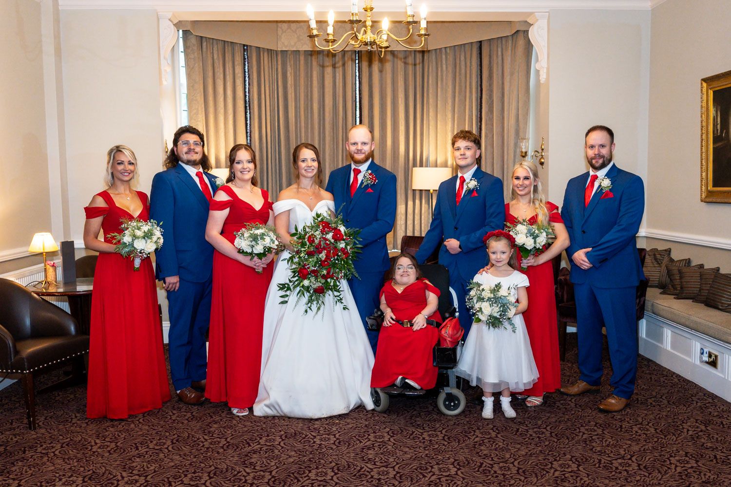 Wedding party in bright red dresses and navy suits poses formally in elegant indoor setting.