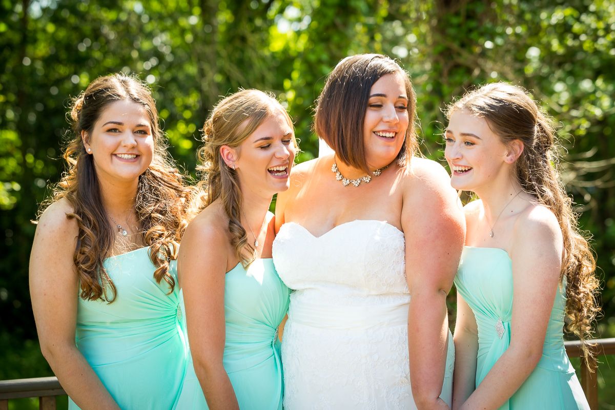 A group of bridesmaids wearing matching mint green dresses laugh together with the bride in white on a sunny day.